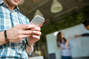 Closeup of hands of young man in checkered shirt using mobile phone while his partners arguing
