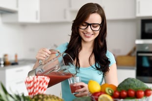 Happy healthy young woman wearing glasses pouring vegetable smoothies freshly made from assorted vegetable ingredients on her kitchen counter